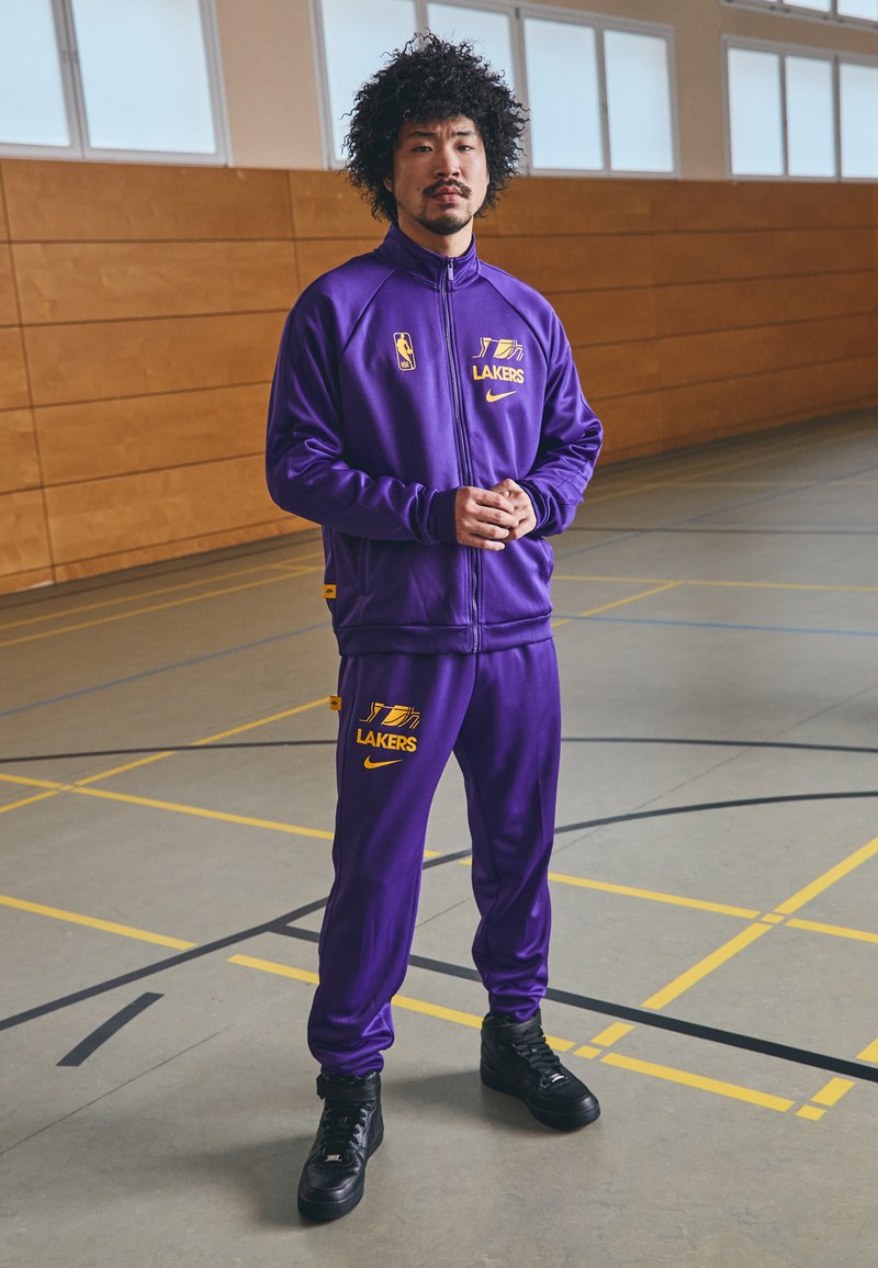 Fato desportivo roxo com o logotipo dos Lakers, casaco com fecho éclair e calças combinando. Ténis pretos; tecido suave e atlético. Ambiente de ginásio com painéis de madeira.