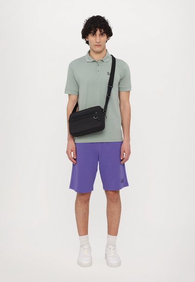 Green polo shirt, purple shorts, and white sneakers. Crossbody bag in textured black with zippers. Simple, modern design.