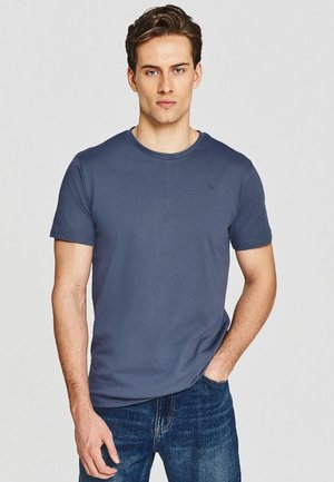 Młody mężczyzna z krótkimi brązowymi włosami, ubrany w gładki niebieski t-shirt i niebieskie dżinsy, stoi z jedną ręką w kieszeni, neutralne tło.