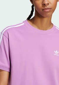 Violetti puuvillapaita lyhyillä hihoilla, ribbauscrew-kaula-aukko ja valkoiset kolme raitaa olkapäillä. Paitassa on pieni valkoinen adidas-logo.