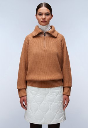 Brauner, gestrickter Pullover mit hohem Kragen und Viertelreißverschluss, kombiniert mit einem weißen, gesteppte Rock mit Rautenmuster und seitlichem Reißverschluss.