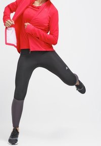 Sportstøj bestående af en lys rød zip-up jakke og sorte leggings med grå detaljer. Leggingsene har en glat og strækbar tekstur.