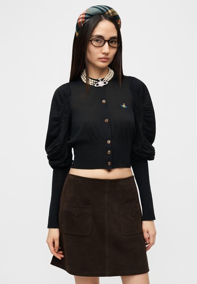 Μαύρη πλεκτή ζακέτα με μανίκια φουσκωτά, κούμπωμα με κουμπιά και μικρό καρφίτσα πλανήτη. Συνδυασμένη με καφέ σουέντ φούστα με τσέπες μπροστά.