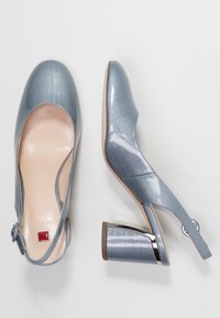 Vaaleansinisiä korkokenkiä, joissa on kiiltävä pinta, pyöreä kärki, nilkkaremmi ja suorakulmainen laatikkokorko, jossa on metallinen yksityiskohta.