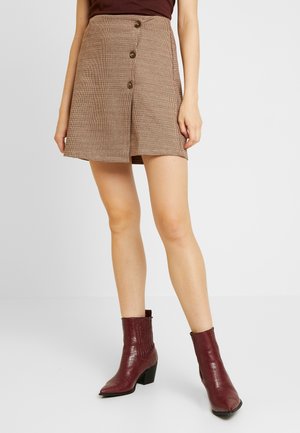 Femme portant une mini-jupe boutonnée devant marron à motifs, associée à des bottines à talons bordeaux, sur fond blanc.