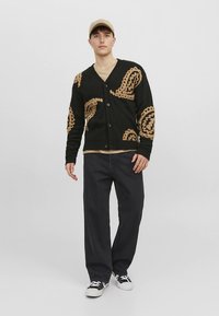 Mustast lõngast kootud jakk, millel on beež paisleymuster, V-kaelus ja nööpidega kinnis, sobitatud mustade pükste ja beeži nokamütsiga.