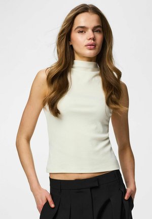 Kvinde med langt bølget brunt hår iført en ærmeløs hvid top med høj krave og sorte bukser, stående med hænderne i lommerne mod en ensfarvet baggrund.