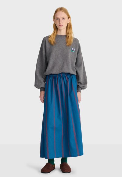 Jeune femme aux longs cheveux roux portant un pull gris oversize, une longue jupe bleue rayée, des chaussettes vertes et des chaussures marron faciles à enfiler, debout sur un fond uni.