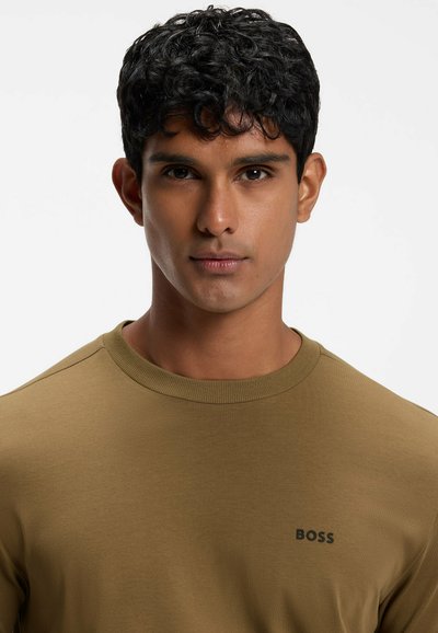 Jeune homme aux cheveux noirs courts et bouclés portant une chemise marron BOSS, faisant face avec une expression neutre sur un fond clair.