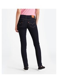 Persona vestida con jeans ajustados oscuros, blusa rosa y zapatillas negras, de pie de espaldas sobre un fondo blanco liso.