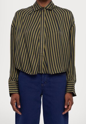 Personne portant une chemise à manches longues avec col, rayures verticales vertes et noires, rentrée dans un jean bleu foncé.