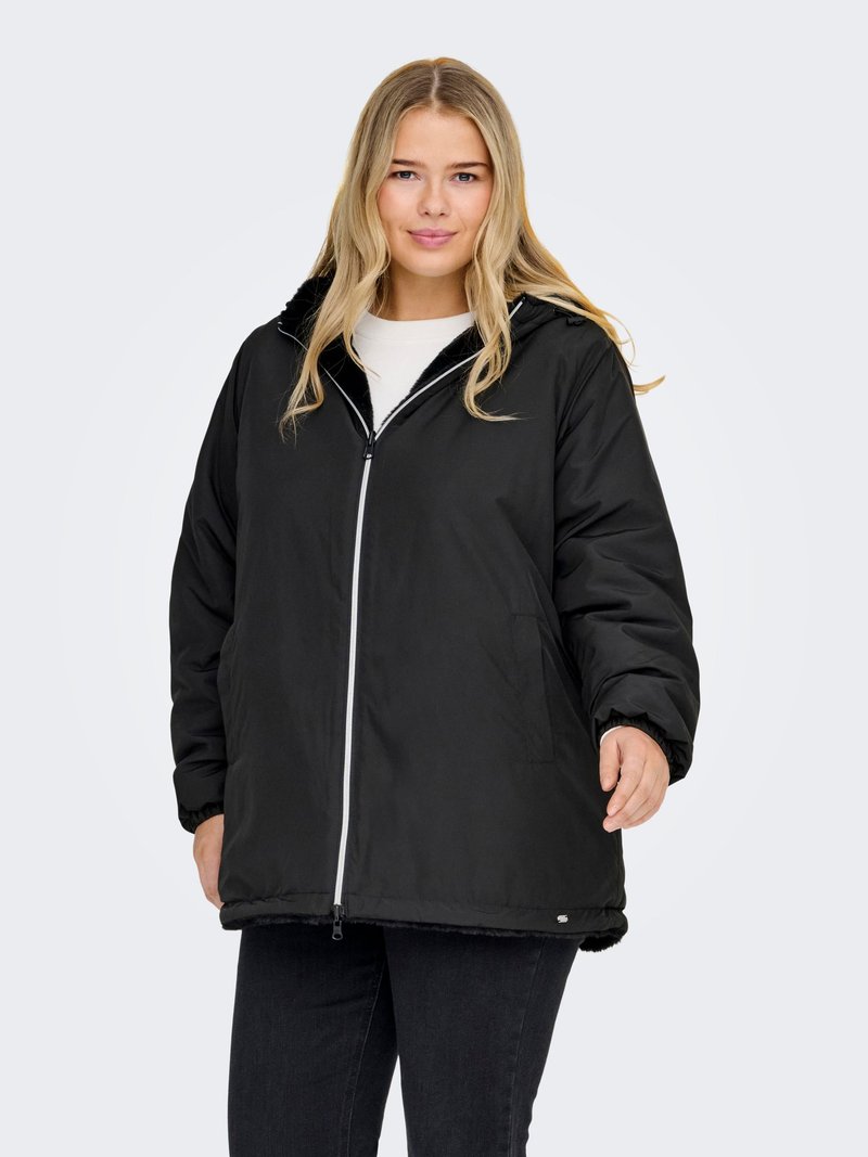 Young woman wearing a black hooded winter jacket with a white zipper, standing against a plain light gray background.