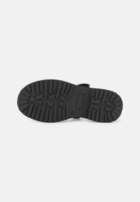 Suela de zapato negra con un patrón de textura para tracción, hecha de goma durable, con un parche rectangular de marca en el centro.