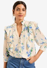 Blusa floreale in beige chiaro con stampe floreali blu e colletto arricciato. Realizzata in tessuto testurizzato con motivi a pois e chiusura a bottoni.