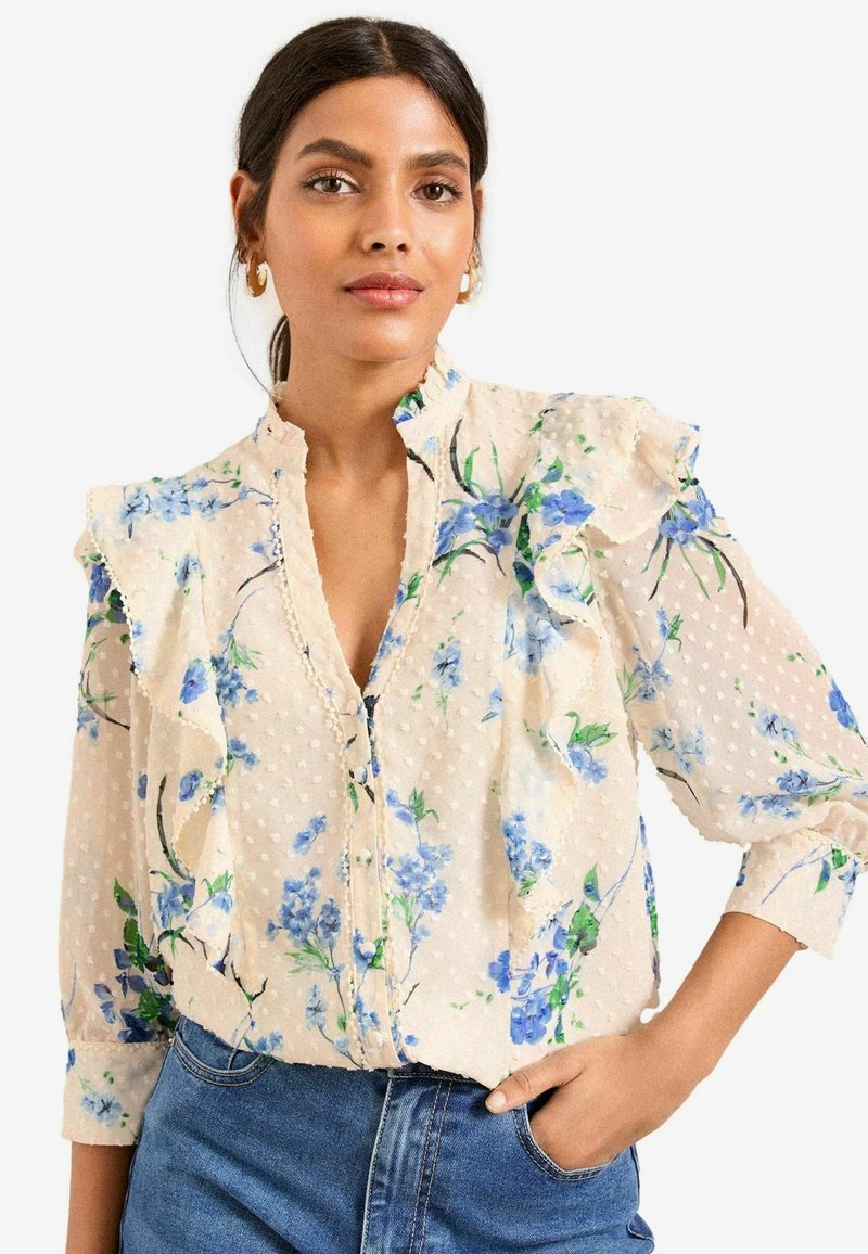 Blusa floreale in beige chiaro con stampe floreali blu e colletto arricciato. Realizzata in tessuto testurizzato con motivi a pois e chiusura a bottoni.