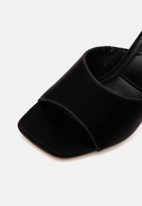 Sandale mule noire à bout ouvert avec une large bride avant et un bout carré sur fond blanc.