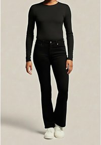 Personne portant un haut noir ajusté à manches longues et un pantalon évasé noir taille haute avec des baskets blanches, debout devant un fond uni.