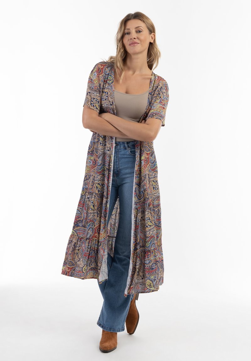 Długi, swobodny kimono z wzorzystego, lekkiego materiału z deseniem paisley w wielu kolorach, zestawiony z beżową bluzką i niebieskimi dżinsami.