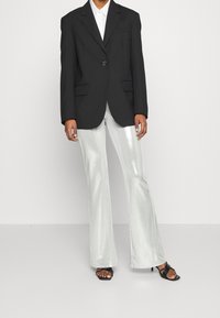 Person wearing black blazer, white shirt, shiny silver flared pants, and black heeled sandals standing on a plain background.