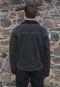 Črna denimska jakna z ovratnikom iz ovčje kože, z vidnimi šivi, stranskimi žepi in rahlo obrabljenim videzom. Obuta s črnimi hlačami.