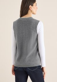 Gilet di maglia grigia con motivo strutturato, dotato di scollo rotondo. Indossato sopra una camicia bianca a maniche lunghe, abbinato a jeans blu scuro.