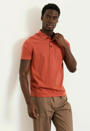 Junger Mann, der den Kragen eines orangen Kurzarm-Poloshirts richtet, trägt hellbraune Hose und steht vor weißem Hintergrund.
