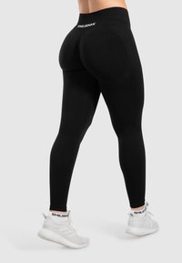 Leggings neri con vita alta, texture liscia e logo prominente sul retro. Indossati con scarpe da ginnastica bianche dal design elevato.