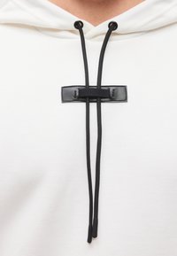 Λευκό φούτερ με κουκούλα με μαύρο δερμάτινο έμπλαστρο που συγκρατεί δύο μαύρα κορδόνια δεμένα στο κέντρο μπροστά, κάτω από τη λαιμόκοψη.