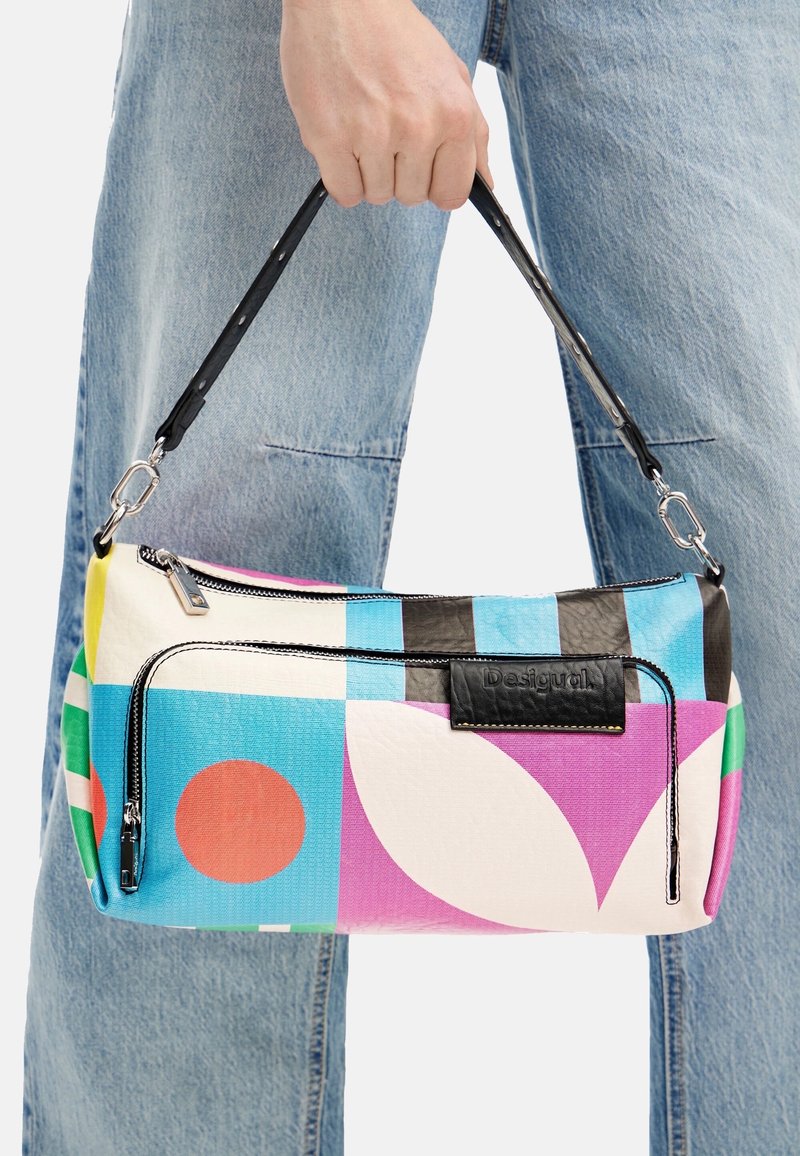 Main tenant un sac à bandoulière rectangulaire coloré avec des motifs géométriques, une sangle noire et une poche zippée à l'avant sur un fond de jean bleu.