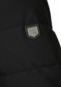 Melnā pūkainā jaka ar šuviem. Iekļauj sešstūra logotipa birku ar metāliskām akcentiem, apzīmētu "MNT 97".