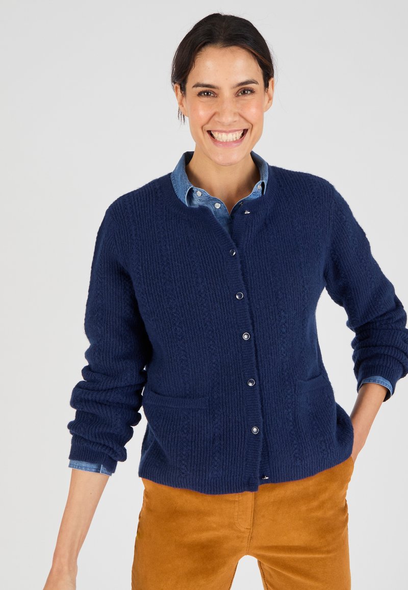 Cardigan lavorato a maglia blu scuro con texture a coste, bottoni frontali e due tasche frontali, indossato sopra una camicia azzurro chiaro con colletto.