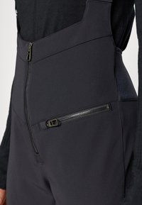 Čierne softshellové nohavice majú diagonálnu zipsovú vrecko, elegantný dizajn a hladkú textúru s minimálnymi švovými detailmi.