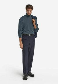 Niebieski sweter z golfem, dopasowane granatowe spodnie, czarny pasek i mokasyny. Model trzyma granatową marynarkę zarzuconą na jedno ramię. Gładkie tło.