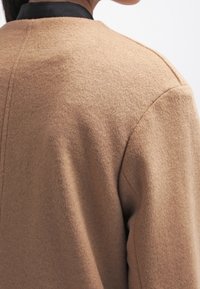 Zbliżenie osoby w beżowym wełnianym płaszczu z czarnym kołnierzem, ukazujące szczegóły pleców i ramion.