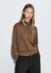 Maglione marrone con collo alla coreana, caratterizzato da bottoni neri sul davanti e una vestibilità comoda. Texture morbida, maniche lunghe, design minimal.