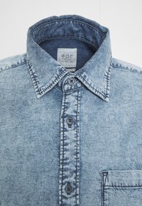 Camisa de mezclilla azul claro con efecto desgastado, cuello puntiagudo, botones negros en la parte delantera y un bolsillo en el pecho del lado derecho.