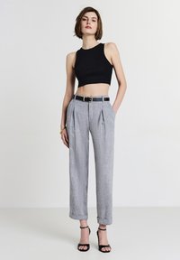 Donna che indossa un top nero a maniche corte, pantaloni grigi a vita alta e plissettati con cintura nera, sandali neri con tacco alto, in piedi davanti a uno sfondo bianco.