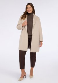 Cappotto in lana beige chiaro con vestibilità sartoriale, design monopetto, ampi revers, due bottoni, abbinato a pantaloni marrone scuro e tacchi chiari.