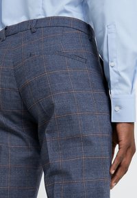 Man i blårutiga byxor och ljusblå skjorta, visar sin nedre rygg och har höger hand vid sidan.