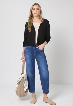 Frau steht und trägt eine schwarze Strickjacke, beige Oberteil, blaue Jeans, beige Slip-On-Schuhe und hält eine braune Tasche mit weiß gemustertem Tuch vor weißem Hintergrund.