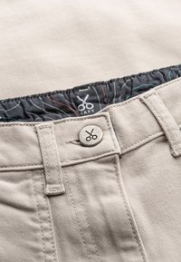 Khaki broek gemaakt van stevig materiaal, met een zilveren knoopsluiting en een gedessineerde tailleband. De binnenvoering heeft rode en groene accenten.