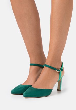 Chaussures à talons hauts en suede vert avec un bout pointu et une sangle à la cheville, présentant un accent vert contrastant sur le talon.
