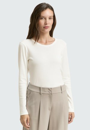 Donna con capelli lunghi e castani, vestita con una maglietta bianca a maniche lunghe e pantaloni beige a vita alta, su uno sfondo chiaro e semplice.