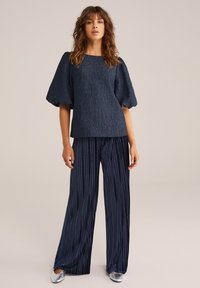 Jūras spēku zila teksturēta blūze ar burbuļveida piedurknēm, kombinēta ar plašām ziliem izšūtām biksēm. Sudraba zābaki noslēdz apģērbu. Vienkāršs, elegants dizains.