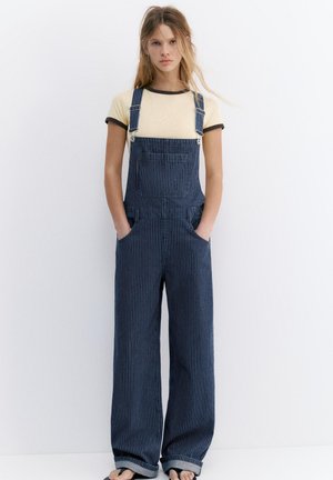 Mladá žena s dlhými vlasmi, ktorá má na sebe béžové tričko a široké námornícky modré nohavice s tenkými pásikmi, stojí s rukami v vreckách na bielom pozadí.