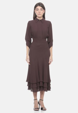 Donna che indossa un abito midi marrone scuro a maniche lunghe con orlo a balze sovrapposte e tacchi alti neri con cinturino alla caviglia, in piedi davanti a uno sfondo semplice.