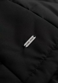 Tessuto di giacca a vento nera con texture trapuntata; presenta un'etichetta argentata con la scritta "OMBRE EXIMIO." Dettagli chiave includono cuciture e superficie liscia.