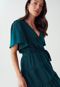 Tafelfarbene Wickelkleid mit kurzen, flatternden Ärmeln, einer taillierten Taille und einem mehrschichtigen Rüschensaum aus weich fallendem Material. Beinhaltet ein gebundenes Detail an der Taille.