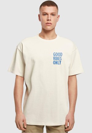 Tricou oversized crem din bumbac, cu mâneci scurte. Prezintă textul albastru „GOOD VIBES ONLY” în zona stângă a pieptului.