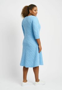 Helles blaues Kleid mit einer strukturierten, wellenförmigen Gestaltung, Dreiviertelärmeln und einem ausgestellten Saum. Tragen mit weißen Turnschuhen.
